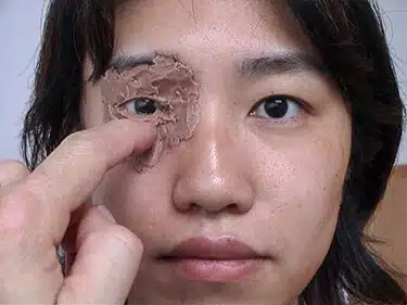 A woman holding up her eye with a piece of cloth in front of it.
